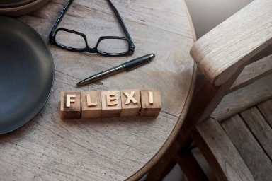 Auf einem Tisch liegen eine Brille und ein Kugelschreiber und aus Holzwürfeln wird das Wort "flexi" gebildet.