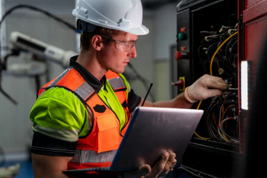 Eine Person mit Helm, Schutzbrille und Computer arbeitet an einer Schalttafel.