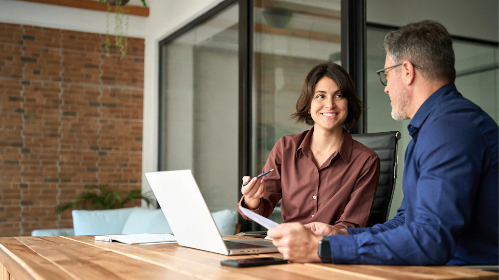 A woman and a man speaking in front of a computer in a meeting room.