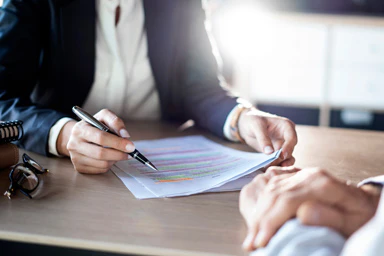 Two people at a desk, one of whom is consulting a document.