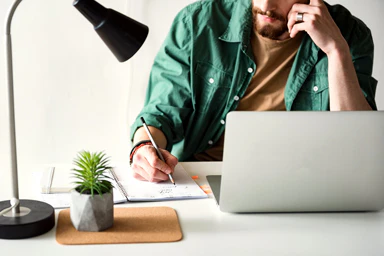 A man in front of his computer takes notes in a notebook.