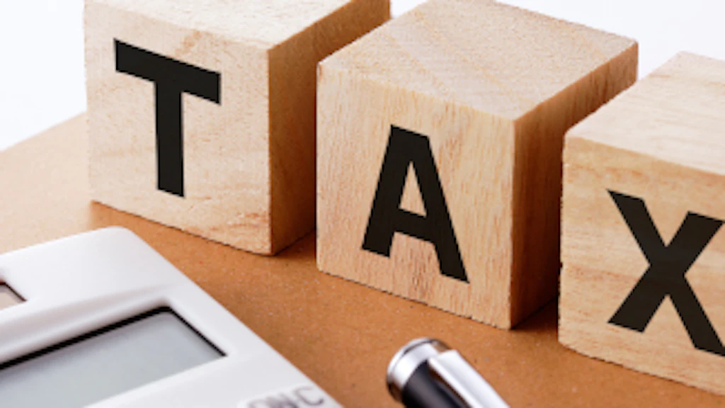 Three wooden blocks marked with the letters t, a and x are placed on a notebook. In the foreground are a calculator and a pen