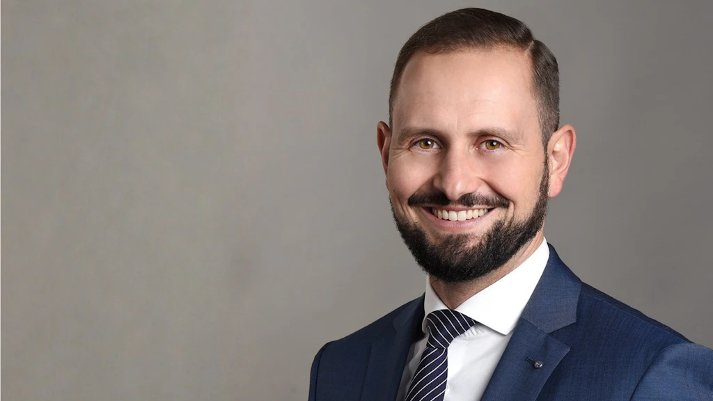 Nicola Tettamanti, blazer bleu foncé, cravate rayée, homme à barbe souriant.