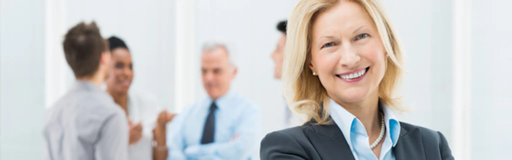 Primo piano di una donna sorridente in abito da ufficio. Alle sue spalle alcuni colleghi, uomini e donne, chiacchierano tra loro.