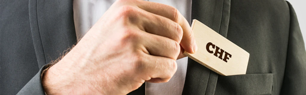 Primo piano sulla mano di un uomo in abiti da ufficio che estrae dal taschino della giacca una tessera di legno sulla quale è stampata la scritta CHF.